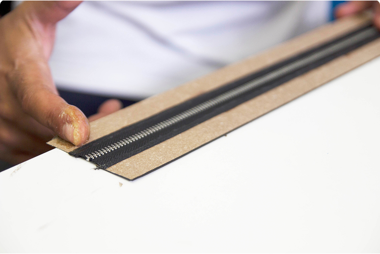 Close-up of a craftsman assembling a zipper onto a leather strip using a wooden base.