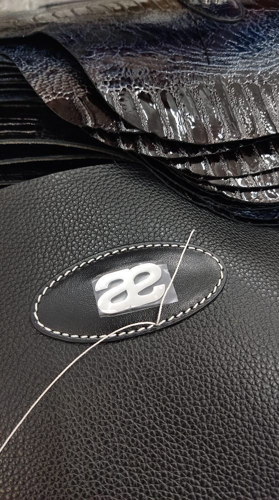 Detail of stitching a silver logo emblem onto black textured leather.